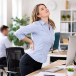 Frau steht im Büro vom Schreibtisch auf und streckt den Rücken zur Entlastung nach langem Sitzen