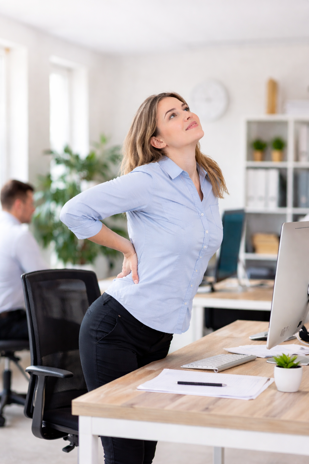 Frau steht im Büro vom Schreibtisch auf und streckt den Rücken zur Entlastung nach langem Sitzen